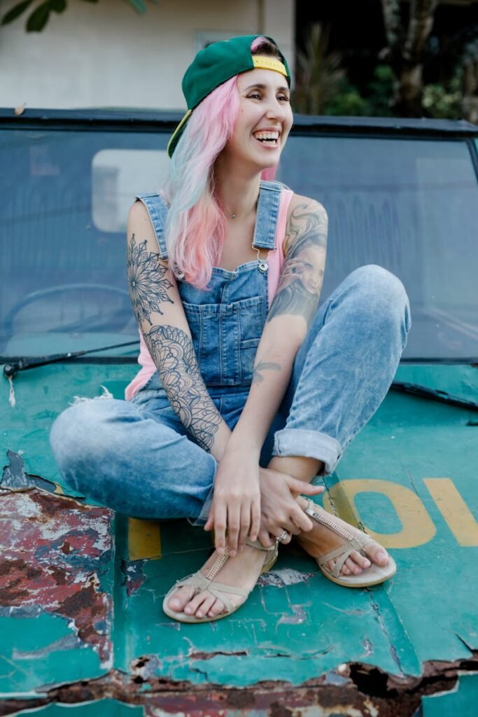 Full body smiling informal female with dyed pink hair and tattooed body wearing denim jumpsuit and cap sitting with legs crossed on weathered rusted car hood and looking away happily